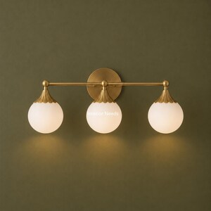 Puede incluir: Un aplique de pared de tres luces con una barra horizontal de lat&oacute;n montada en una placa circular. Cada luz consta de un globo esf&eacute;rico de vidrio esmerilado blanco unido a una tapa de lat&oacute;n festoneada. El texto Interior Needs es visible.