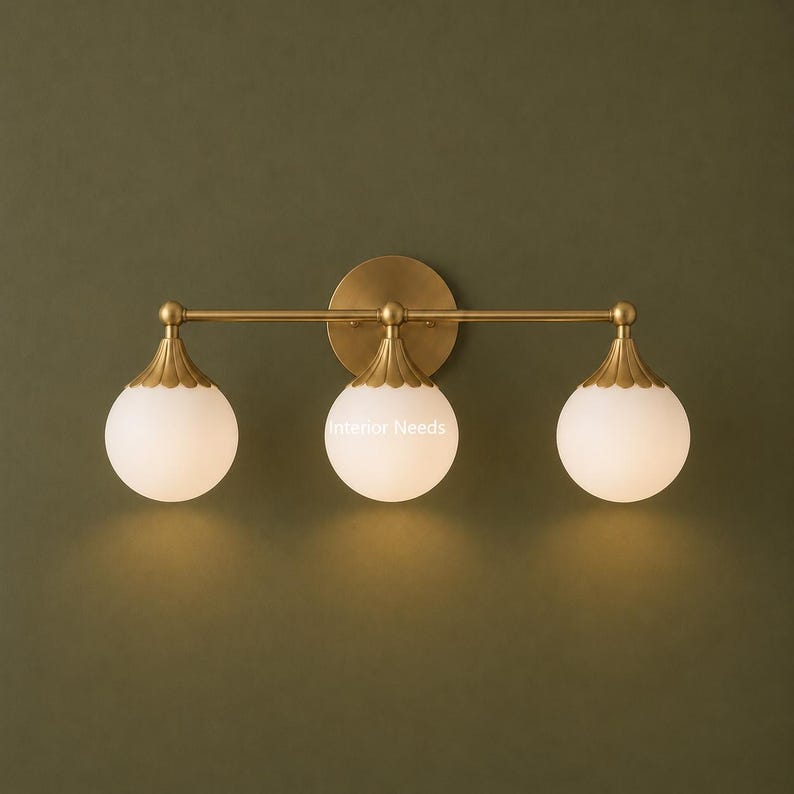 Puede incluir: Un aplique de pared de tres luces con una barra horizontal de lat&oacute;n montada en una placa circular. Cada luz consta de un globo esf&eacute;rico de vidrio esmerilado blanco unido a una tapa de lat&oacute;n festoneada. El texto Interior Needs es visible.