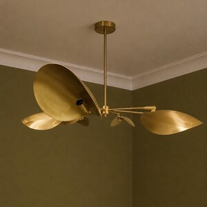 May include: A brass chandelier featuring multiple curved, leaf-shaped metal shades extending from a central rod. The fixture has a metallic gold finish and includes exposed light bulb sockets positioned within the concave surfaces of the shades.