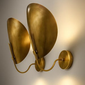 May include: A pair of brass wall sconces with a warm, golden finish. Each sconce features a curved, leaf-shaped shade and a circular base. The design includes a curved arm connecting the shade to the base, creating a modern, elegant look.