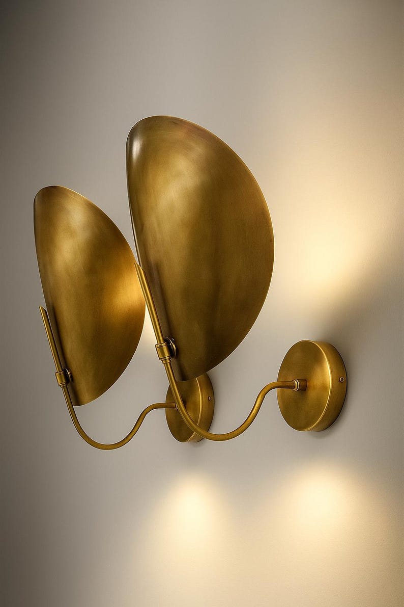 May include: A pair of brass wall sconces with a warm, golden finish. Each sconce features a curved, leaf-shaped shade and a circular base. The design includes a curved arm connecting the shade to the base, creating a modern, elegant look.