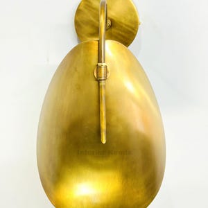 May include: A brass wall sconce with a teardrop-shaped shade and a circular backplate. The sconce has a curved arm and a vertical support. The text "Interior Needs" is visible on the shade.