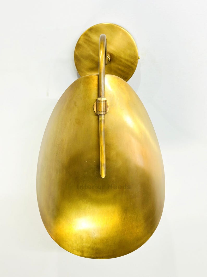 May include: A brass wall sconce with a teardrop-shaped shade and a circular backplate. The sconce has a curved arm and a vertical support. The text "Interior Needs" is visible on the shade.
