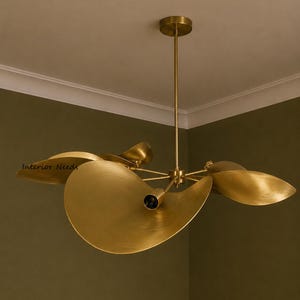 May include: A brass chandelier featuring multiple curved, petal-shaped shades extending from a central rod. The fixture has a brushed metallic finish and includes a small dark bulb socket at the center of each shade. The text Interior Needs is visible.