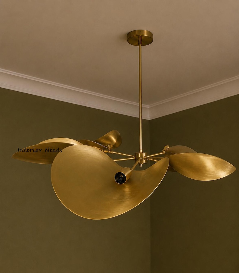 May include: A brass chandelier featuring multiple curved, petal-shaped shades extending from a central rod. The fixture has a brushed metallic finish and includes a small dark bulb socket at the center of each shade. The text Interior Needs is visible.
