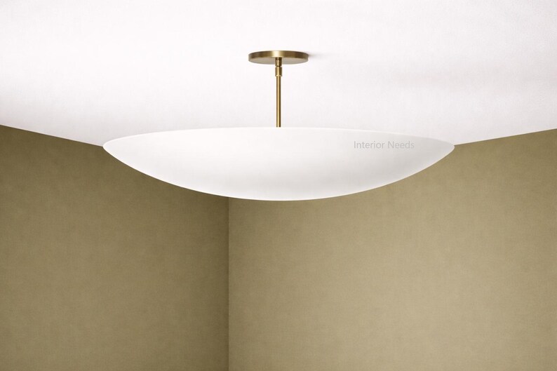 May include: A modern, white, oval-shaped ceiling light fixture with a brass-colored stem and ceiling mount. The fixture is suspended from the ceiling and provides ambient lighting. The text "Interior Needs" is visible on the light.