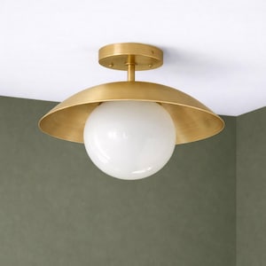 May include: A brass-colored ceiling light fixture with a white globe-shaped light. The fixture has a circular base and a curved shade that surrounds the globe. The light is mounted on a white ceiling.