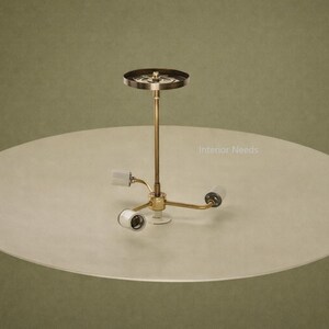 May include: A mid-century modern ceiling light fixture with a brass finish and three light sockets. The fixture is mounted on a large, round, off-white shade. The background is a muted olive green.