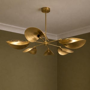 May include: A brass-colored chandelier featuring six curved, leaf-shaped shades extending from a central rod. The fixture is mounted to the ceiling with a circular canopy. The text "Interior Needs" is visible on one of the metal shades.
