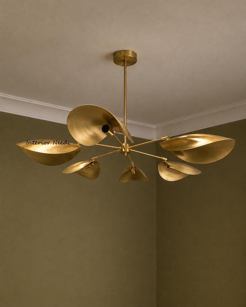 May include: A brass-colored chandelier featuring six curved, leaf-shaped shades extending from a central rod. The fixture is mounted to the ceiling with a circular canopy. The text "Interior Needs" is visible on one of the metal shades.