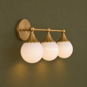 Puede incluir: Un aplique de pared de tres luces con acabado en lat&oacute;n cepillado, un soporte circular y tres globos esf&eacute;ricos de vidrio blanco. Cada globo est&aacute; rematado con una tapa de lat&oacute;n festoneada. El texto Interior Needs es visible en el globo central.