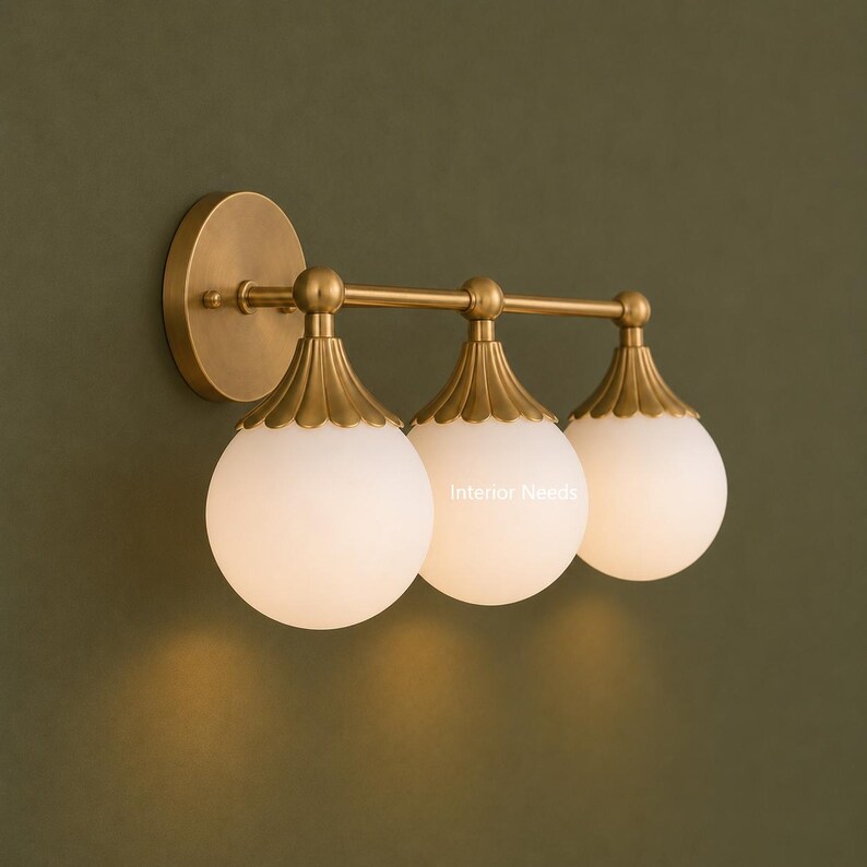 Puede incluir: Un aplique de pared de tres luces con acabado en lat&oacute;n cepillado, un soporte circular y tres globos esf&eacute;ricos de vidrio blanco. Cada globo est&aacute; rematado con una tapa de lat&oacute;n festoneada. El texto Interior Needs es visible en el globo central.