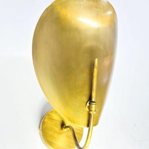 May include: A brass wall sconce with a curved, shell-like shade. The sconce has a circular base and a curved arm that supports the shade. The text "Interior Needs" is visible on the shade. The finish is a warm, golden color.
