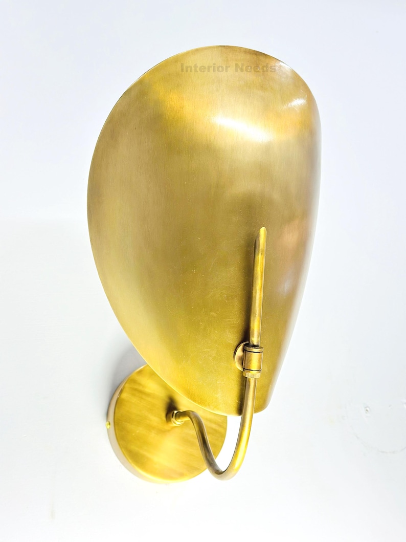 May include: A brass wall sconce with a curved, shell-like shade. The sconce has a circular base and a curved arm that supports the shade. The text "Interior Needs" is visible on the shade. The finish is a warm, golden color.