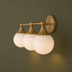 Puede incluir: Un aplique de pared de tres luces con acabado en lat&oacute;n cepillado, una placa circular y tres globes esf&eacute;ricos de vidrio esmerilado blanco. Cada globo tiene una tapa de lat&oacute;n festoneada. El texto Interior Needs es visible en el vidrio.