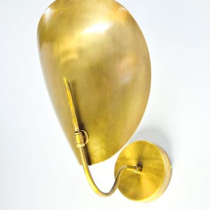 May include: A brass wall sconce with a large, curved shade and a circular base. The sconce has a warm, golden hue and a sleek, modern design. The shade is shaped like a teardrop and is attached to the base with a curved arm.