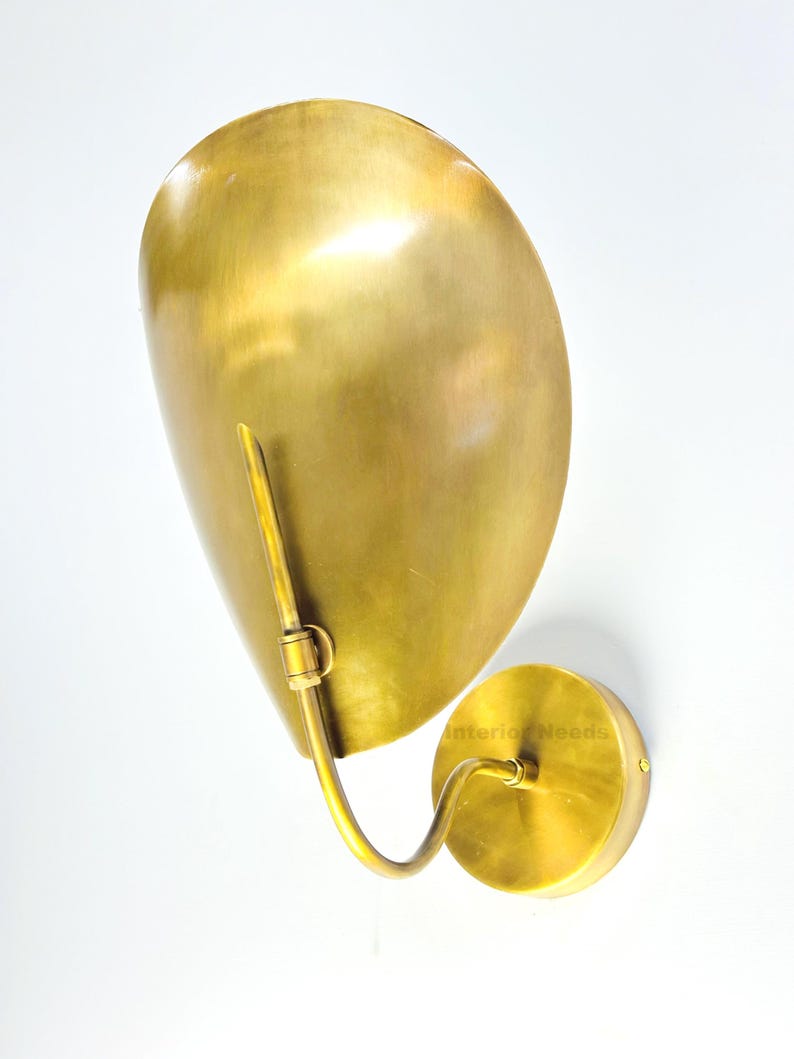 May include: A brass wall sconce with a large, curved shade and a circular base. The sconce has a warm, golden hue and a sleek, modern design. The shade is shaped like a teardrop and is attached to the base with a curved arm.
