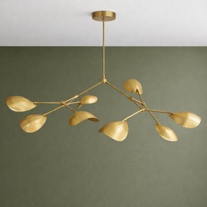 Op de afbeelding: Een moderne kroonluchter in messingkleur, met een takachtig ontwerp. De armatuur heeft meerdere gebogen armen, elk met een bladachtig licht. Het hangt aan een centrale stang.