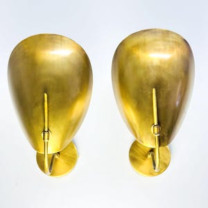 May include: Two gold-colored wall sconces with a teardrop-shaped shade and a curved arm. The sconces have a circular base and a vertical rod extending from the shade. The finish is a shiny, metallic gold.