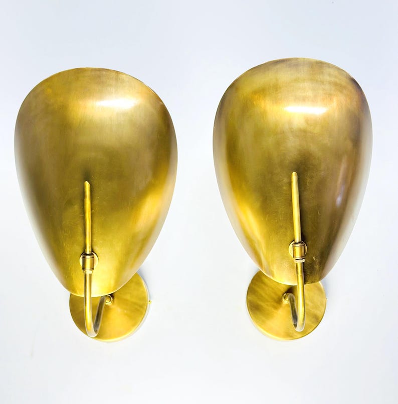 May include: Two gold-colored wall sconces with a teardrop-shaped shade and a curved arm. The sconces have a circular base and a vertical rod extending from the shade. The finish is a shiny, metallic gold.