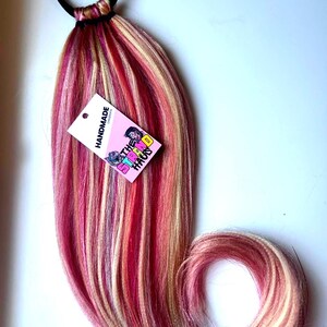 May include: A hair extension piece with a black elastic band, featuring strands of pink, red, and blonde hair. A small tag with the text "HANDMADE" and "THE STIRRED HAUS" is attached.