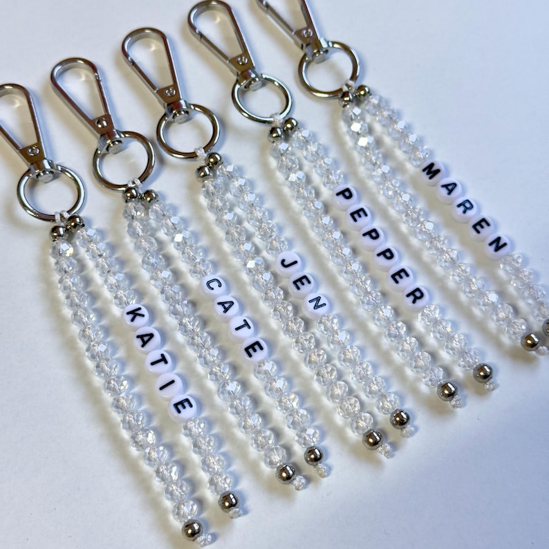 May include: Five keychains with clear crystal beads and silver-tone hardware. Each keychain has a different name spelled out in white letter beads: Katie, Cate, Jen, Pepper, and Maren. The keychains are arranged on a white surface.