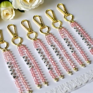 May include: A collection of personalized keychains with gold-colored clasps and pink beaded tassels. Each keychain features a name spelled out in black letters on white beads. The names include Kayla, Abby, Arielle, Casey, Alexa, and Johnette.