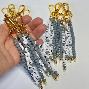 May include: Keychains with gold-colored clasps and beaded strands in gray and white. The keychains spell out "BETA PHI" and "PI BETA PHI" with white letter beads. Gold-colored accents are at the bottom of each strand.