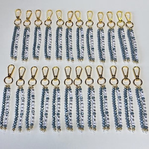 May include: Keychains with gold-colored clasps and beaded tassels in gray and white. The tassels spell out "PI BETA PHI" in white letter beads. The keychains are arranged in rows.