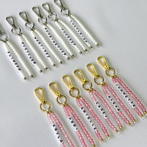 May include: Keychains with names spelled out in white letter beads. The keychains have gold-colored clasps and are strung with white or pink beads. Names include: Puthoff, Fletcher, Martinez, Altstaetter, Youssef, Shayleigh, Kayla, Abby, Arielle, Casey, Alexa, and Johnette.