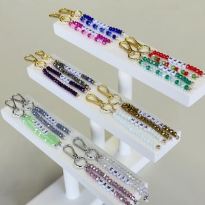 May include: Keychains with beaded strands in various colors, including blue, pink, green, and gold. Each keychain features a name spelled out with white letter beads. Gold and silver-toned clasps are attached.
