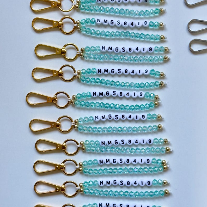 May include: A collection of keychains with gold-colored clasps and light blue beaded accents. Each keychain features a string of light blue beads and a row of white letter beads spelling out "NMG S 8419".