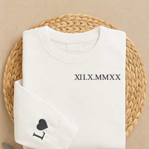 May include: A white sweatshirt with the Roman numeral date XII.X.MMXX printed in black. The left sleeve features a black heart and the letter "L". The sweatshirt is folded on a woven, circular mat.