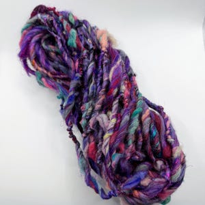 May include: A vibrant skein of hand-spun yarn in shades of purple, pink, teal, and gold. The thick, textured yarn is ideal for fiber art projects, knitting, or crochet. The yarn has a unique, artisanal appearance.