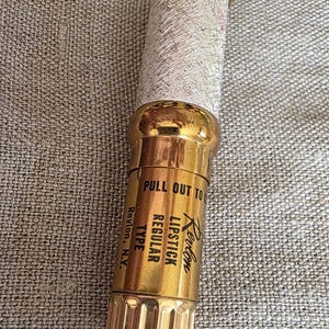 Può includere: Rossetto Revlon vintage in un astuccio dorato e argentato. L'astuccio del rossetto ha un corpo argentato strutturato con dettagli dorati nella parte superiore e inferiore. Il testo "Revlon Lipstick Regular Type" è stampato sull'astuccio. Su una superficie di colore neutro.