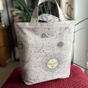 May include: A beige tote bag with a celestial design featuring suns, moons, stars, and zodiac symbols. The bag has two handles and is made of a textured fabric. The bag is sitting on a red surface.