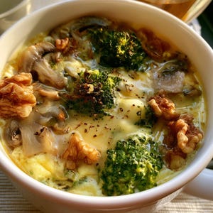 Può includere: Una tazza in ceramica bianca contenente un piatto a base di uova con cimette di broccolo verde brillante, funghi marroni affettati e gherigli di noce interi. La superficie è ricoperta di formaggio fuso con una spolverata di pepe nero.
