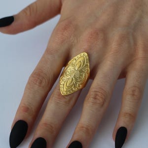 以下が含まれることがあります： 細長い葉の形をしたデザインのゴールドカラーのステートメントリング。リングは複雑な花柄と渦巻き模様が特徴で、詳細で装飾的な外観を作り出しています。リングは指に着用され、そのサイズとデザインを示しています。