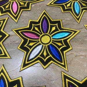 May include: Embroidered star-shaped patches with a black base and gold trim. Each patch features a central flower design with petals in various colours, including pink, blue, purple, and white. The patches are arranged on a neutral surface.