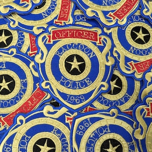 May include: A collection of blue and gold embroidered patches. Each patch features a shield design with the words "Officer," "Raccoon Police," and the year "1969." A black star is in the center of each patch.