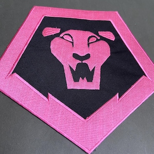 Op de afbeelding: Een roze en zwarte geborduurde patch met een gestileerde leeuwenkop in een vijfhoek. De leeuw is zwart, met roze contouren en details. De patch heeft een roze rand.