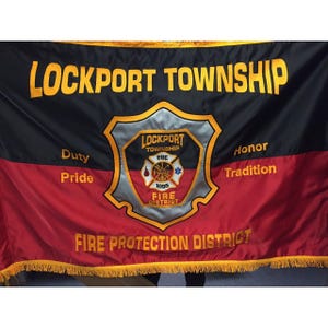 Op de afbeelding: Een zwart, rood en goudkleurige vlag met de woorden "LOCKPORT TOWNSHIP" bovenaan. De vlag heeft een schildembleem met de woorden "Duty", "Pride", "Honor" en "Tradition". De onderkant van de vlag luidt "FIRE PROTECTION DISTRICT".