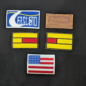 May include: A selection of embroidered patches. One patch displays the text "FAST END" in white on a blue and white background. Another patch reads "frontier" in brown. There are two yellow and red patches, and one American flag patch.