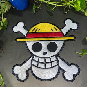 May include: Embroidered patch of the One Piece Jolly Roger. The design features a white skull with black eyes and a stitched smile, crossed bones, and a yellow and red straw hat. The patch has a black border, suitable for application on clothing or accessories.