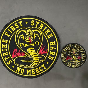 Peut inclure: Deux écussons Cobra Kai. Le plus grand est rond, noir avec du texte jaune et un emblème de cobra jaune. Le texte se lit "Strike First, Strike Hard, No Mercy." Le plus petit écusson a le même design.