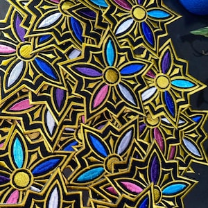 Könnte beinhalten: Eine Ansammlung gestickter Blumenaufnäher. Jeder Aufnäher hat einen schwarzen Hintergrund mit goldenen Konturen und farbigen Blütenblättern in Blau, Lila, Rosa und Weiß. Die Aufnäher haben eine sternartige Form.