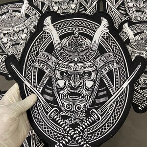 Könnte beinhalten: Schwarz-weißer, gestickter Aufnäher mit einem detaillierten Samurai-Kriegermasken-Design. Der Aufnäher hat einen kreisförmigen Rand mit gekreuzten Schwertern und komplizierten Mustern. Das Design ist kühn und grafisch, für Kleidung.