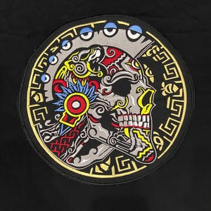 May include: A circular patch with a detailed skull design. The skull is decorated with intricate patterns in red, yellow, blue, and grey, set against a black background. The patch has a gold border and is likely for clothing or accessories.