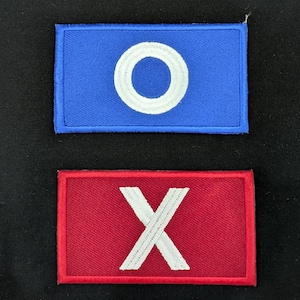 Könnte beinhalten: Zwei rechteckige, gestickte Aufnäher. Der obere ist blau mit einem weißen Kreis. Der untere ist rot mit einem weißen 'X'. Beide haben einen farbigen Rand, wahrscheinlich zur Identifizierung.