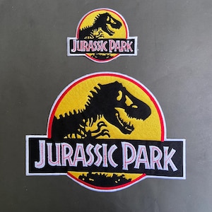 Pode incluir: Dois patches bordados com o logotipo "Jurassic Park". O patch maior apresenta uma silhueta de dinossauro preta em um círculo amarelo, com o texto "JURASSIC PARK" abaixo. O patch menor tem o mesmo design.